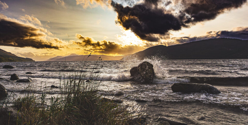 Sonnenuntergang über einem stürmischen See mit dunklen Wolken, Bergen im Hintergrund und Wellen, die gegen Felsen schlagen.
