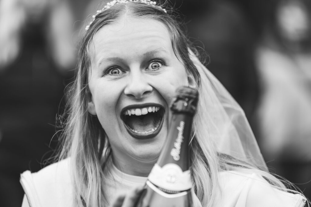 Lachende Frau im Brautschleier hält Sektflasche in der Hand, Gesichtsausdruck voller Freude und Überraschung, Nahaufnahme.