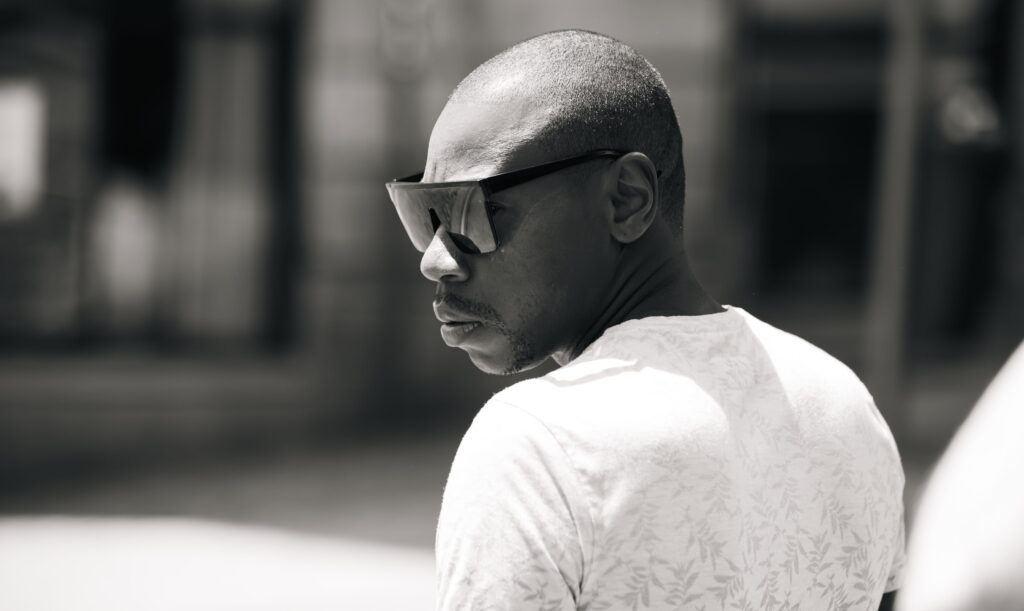 Mann mit rasiertem Kopf und großer Sonnenbrille blickt ernsthaft über die Schulter, aufgenommen in Schwarz-Weiß.