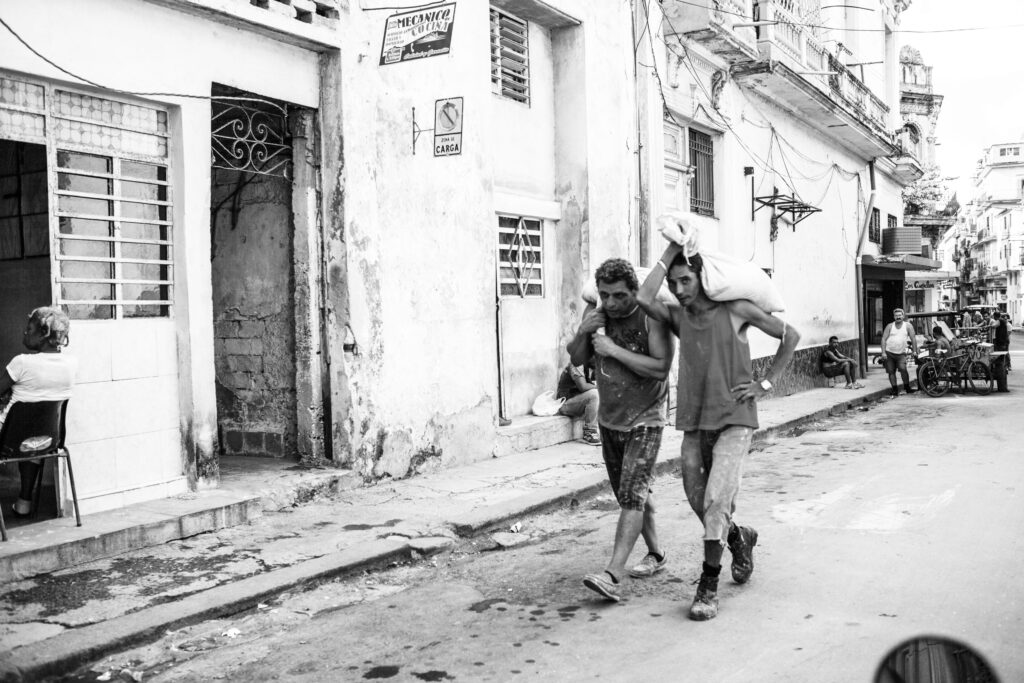 Zwei Männer tragen gemeinsam einen großen Sack durch eine Straße in Havanna, Kuba; eine Frau sitzt am offenen Hauseingang.