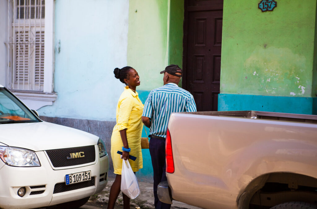 Lächelnde Frau in gelbem Kleid schaut alten Mann an, eingerahmt von zwei Pick-ups in Havanna.