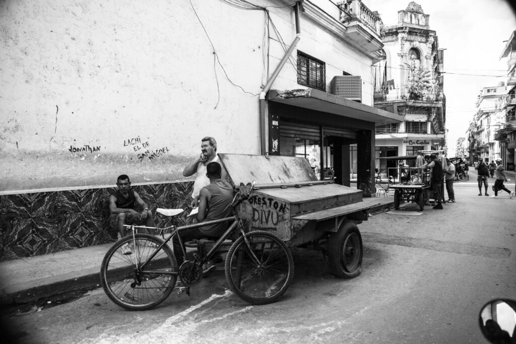 Drei Männer unterhalten sich neben einem Fahrradanhänger in einer Straße in Havanna, Kuba; weitere Menschen im Hintergrund sichtbar.