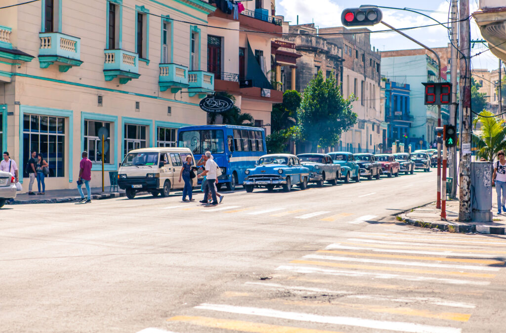 Kreuzung in Havanna mit gehenden Passanten und bunten Oldtimern im Hintergrund.