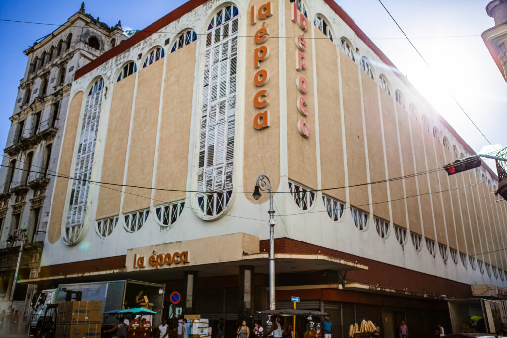 Hinter dem Einkaufszentrum in Havanna spiegelt sich die Sonne im Himmel, LKW wird entleert, Menschen laufen vorbei.