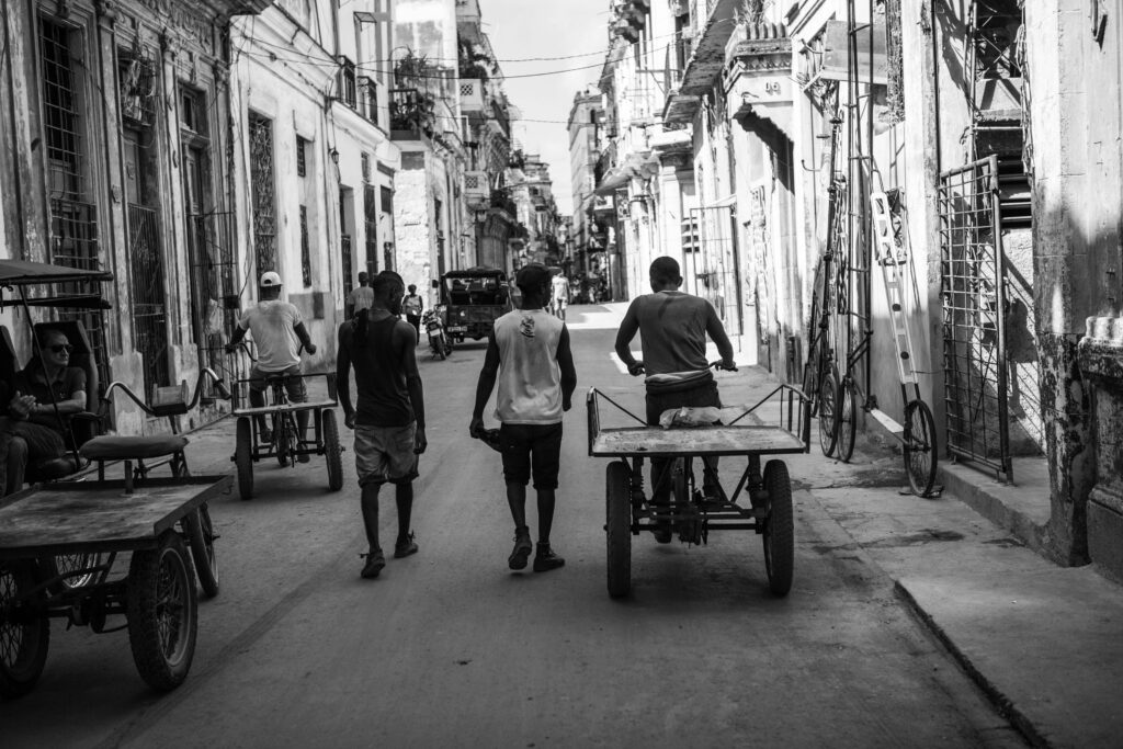 Schwarzweiß-Foto von Männern mit Handkarren und Fahrradrikschas in einer engen Straße der Altstadt von Havanna, Kuba.