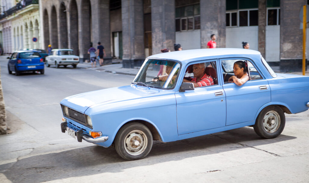 Blaues Auto mit drei Personen fährt durch eine Straße in Havanna, Kuba; koloniale Architektur und Passanten im Hintergrund.