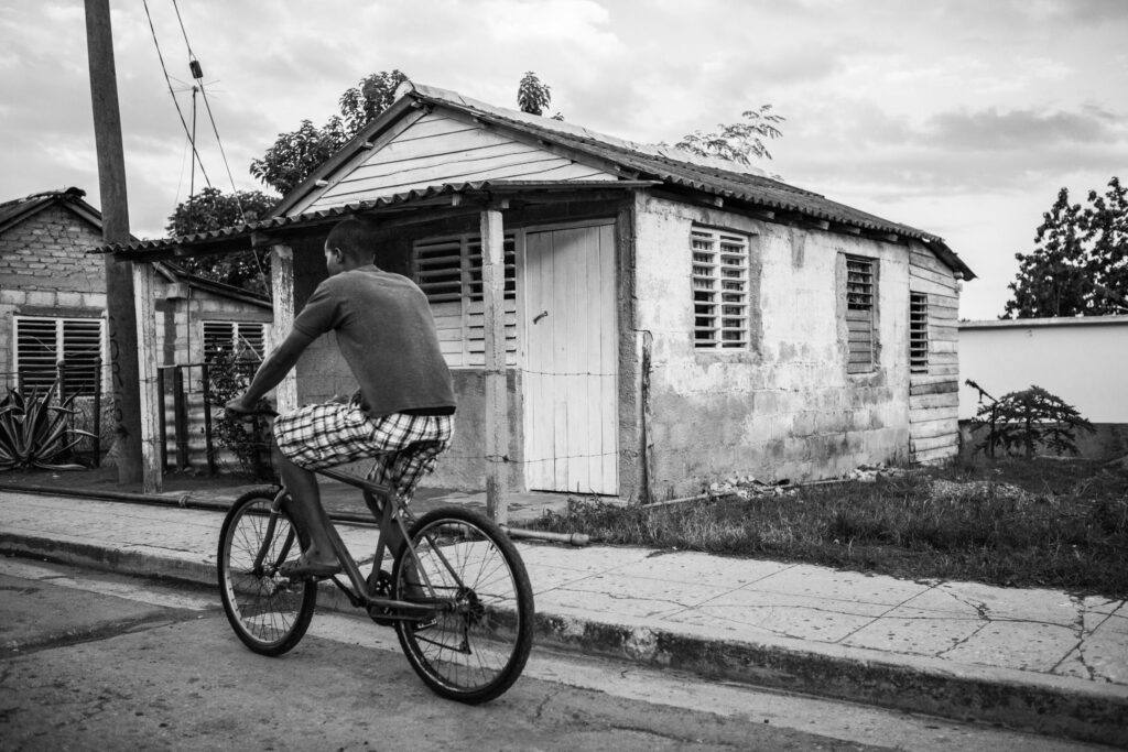 Mann mit Fahrrad fährt barfuß an einfachen Häusern in Kuba vorbei; Szene bei bewölktem Himmel.