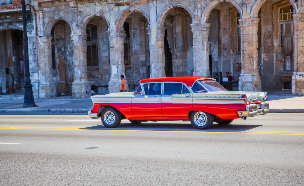 Oldtimer fährt am Malecón in Havanna vorbei, Straßenseite mit DJ, koloniales Haus mit großen Bogenportalen und farbiger Fassade.