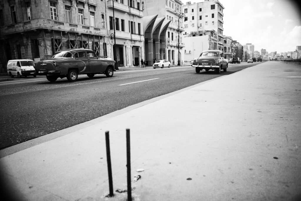 Oldtimer fahren auf einer breiten Straße entlang des Malecón in Havanna, Kuba, mit historischen Gebäuden im Hintergrund.