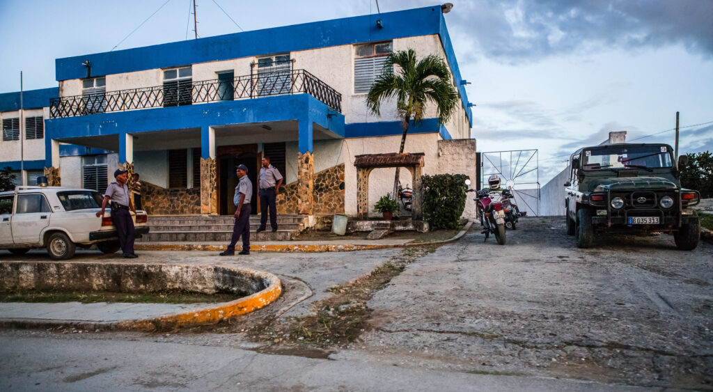 Drei uniformierte Polizisten stehen vor ihrer Wache, einem historischen Haus in Kuba, einer lehnt am Auto.