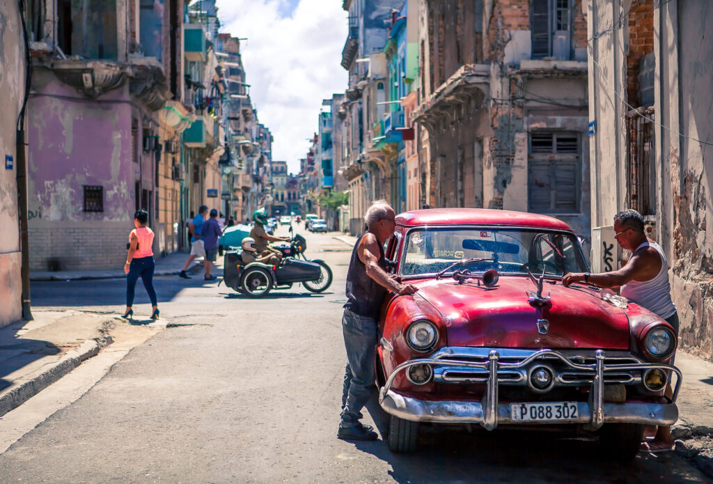 Zwei Männer stehen an einem Oldtimer im Vordergrund in Havanna, Kuba; koloniale Gebäude und Passanten im Hintergrund.