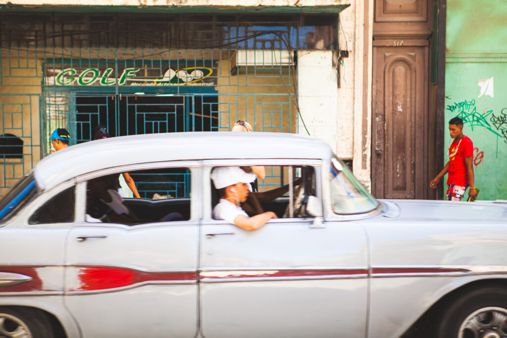 Alter weißer Oldtimer fährt durch eine Straße in Havanna, junger Mann mit Kappe schaut aus dem Fenster.
