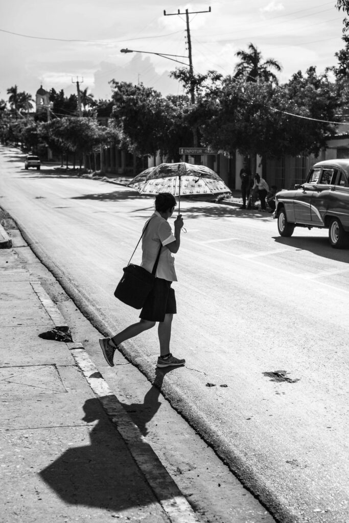 Frau mit Regenschirm überquert bei Sonne eine Straße in Kuba, ein Oldtimer und Schatten im Bild sichtbar.