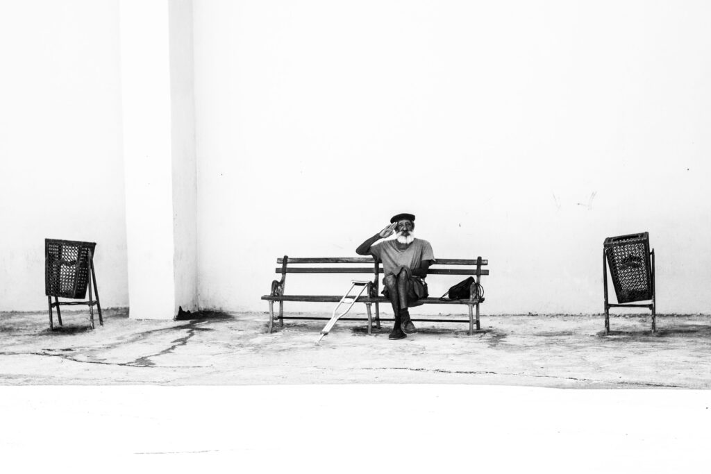 Älterer Mann mit Krücke sitzt auf Bank vor weißer Wand, hebt salutierend die Hand, Mülleimer links und rechts im Bild.